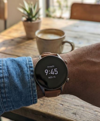 A close-up photograph of an Amazfit GTR 3 Pro smartwatch worn on a wrist with a brown leather strap. The digital watch face displays the time "9:45" and date "WED 15". The person wears a blue denim shirt cuff, resting their arm on a rustic wooden table next to a ceramic coffee mug and a small potted plant in a sunlit cafe setting.